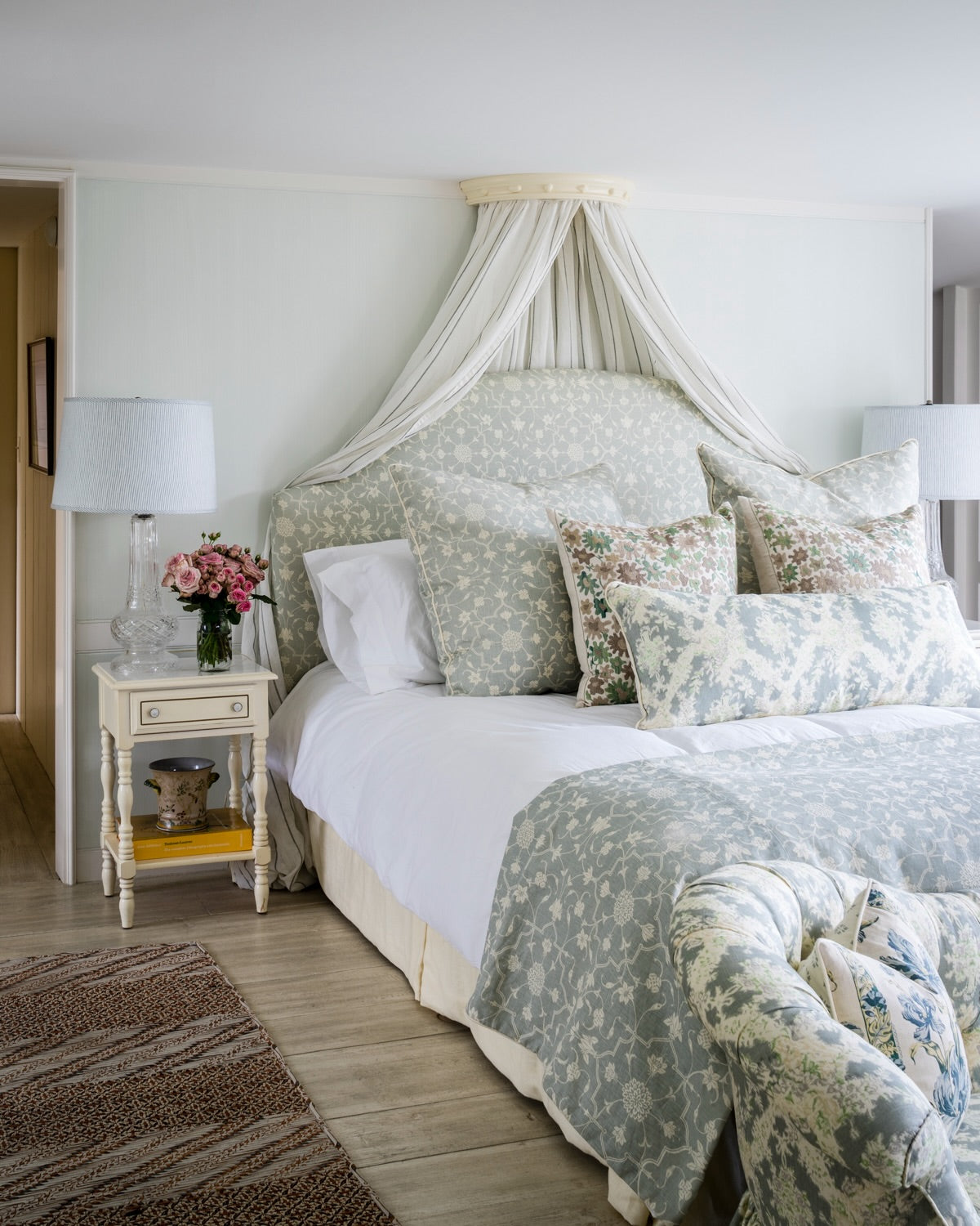 A beautifully styled bedroom featuring a bed with a large upholstered headboard, matching comforter, and pillows in Abu Fog. The fabric canopy is in Sheer Stripe Blue and the setee and lumbar pillow is in Ikat Blue. The bedroom is designed by Kathryn M. Ireland.