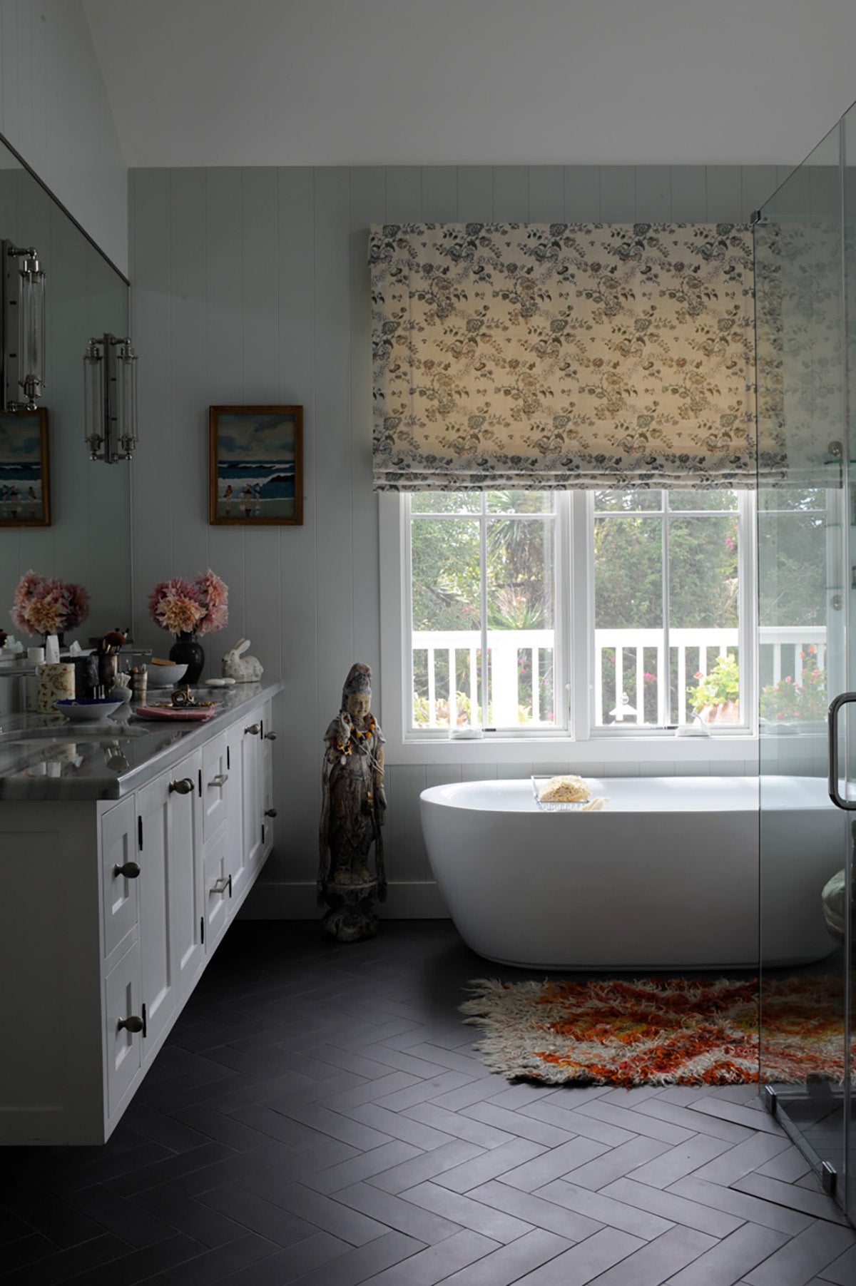 A modern bathroom interior with a freestanding white bathtub positioned by a large window with a Bouquet Blue roman shade, designed by Kathryn M. Ireland