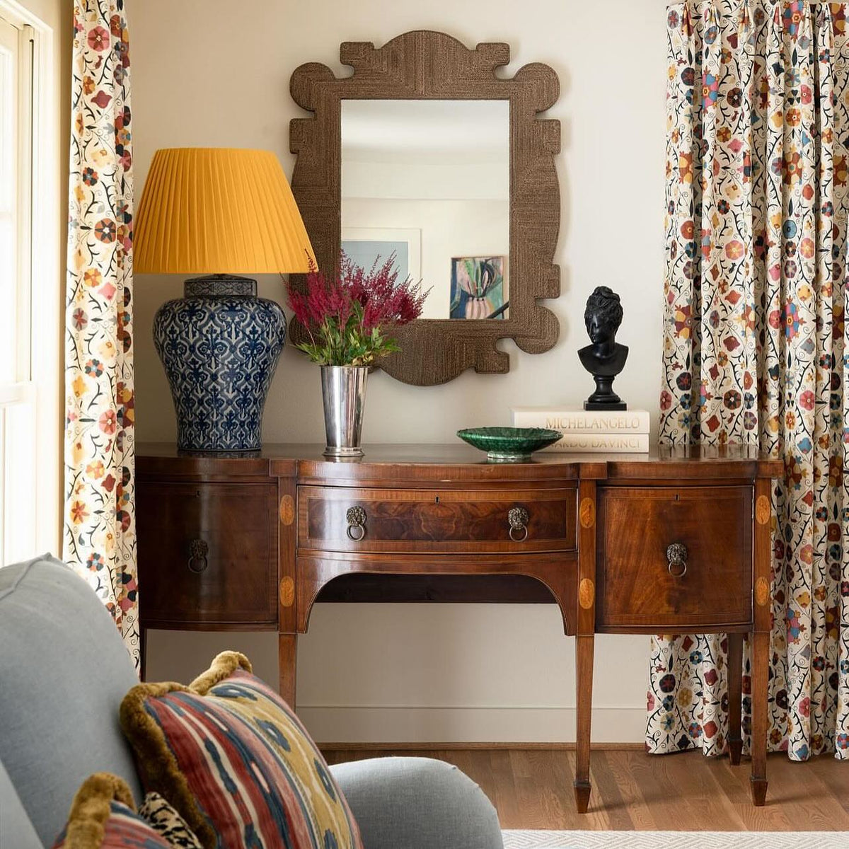 An interior scene featuring a dark wood console table beneath an ornate wooden mirror, with a patterned lamp, and Safi Suzani Indigo Passion curtains, designed by Kathryn M. Ireland.