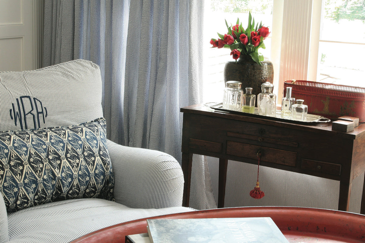 A cozy living room corner featuring a light gray ribbed armchair with a Diamond Batik Indigo accent pillow and a dark wood console table holding a vase of red tulips and glassware designed by Kathryn M. Ireland