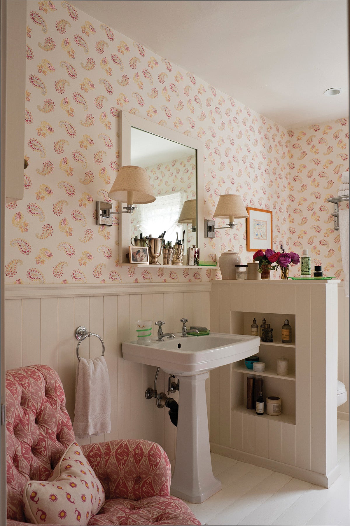A styled bathroom interior featuring a white pedestal sink under a wall mirror, set against Quilt wallpaper, and a matching Diamond Batik Red armchair designed by Kathryn M. Ireland