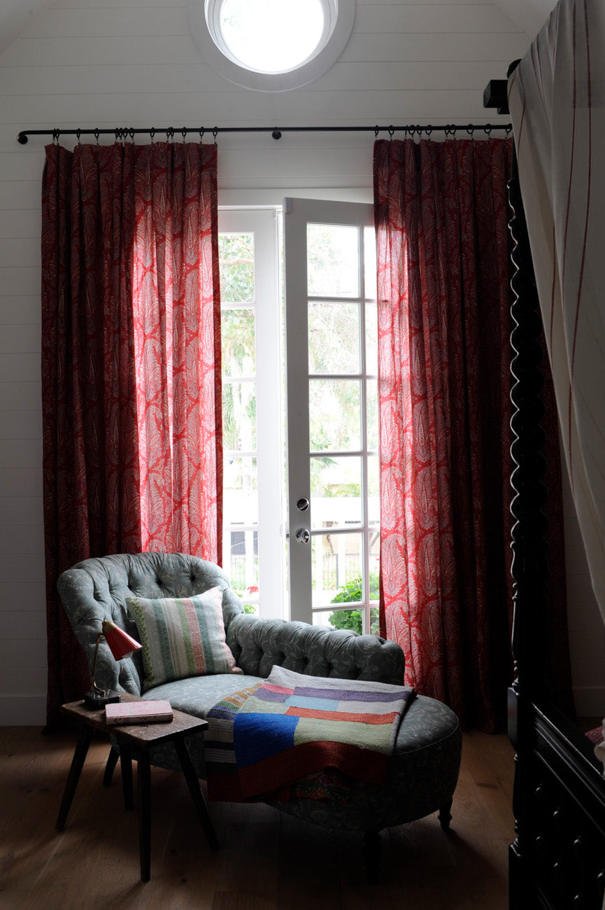 A styled interior scene with a window dressed in Egerton Paisley Red curtains, with a gray upholstered armchair and chaise with a Keltic Stripe Turquoise pillow, designed by Kathryn M. Ireland