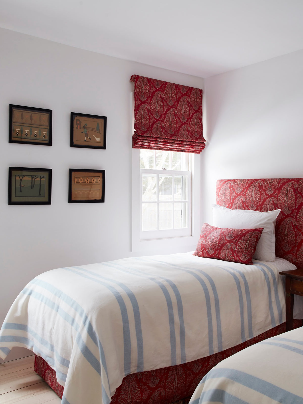 A bedroom featuring twin beds with a headboard, pillow, and bed curtain in Egerton Paisley Red with a matching roman shade, designed by Kathryn M. Ireland.