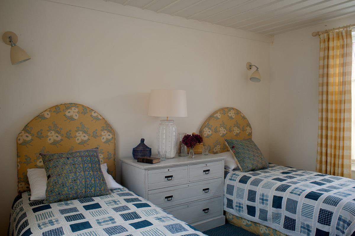 A twin bedroom setup with two single beds featuring Floral Batik Yellow headboards, patterned bedding, a nightstand with a lamp, and Ikat Check Yellow curtains, designed by Kathryn M. Ireland