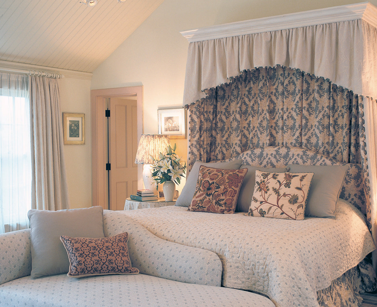 A classic bedroom scene featuring a four-poster canopy bed with an Ikat Blue headboard and matching drapes, dressed in Floral Blue bedding and a Floral Blue upholstered setee, with Sheer Stripe Blue curtains, designed by Kathryn M. Ireland