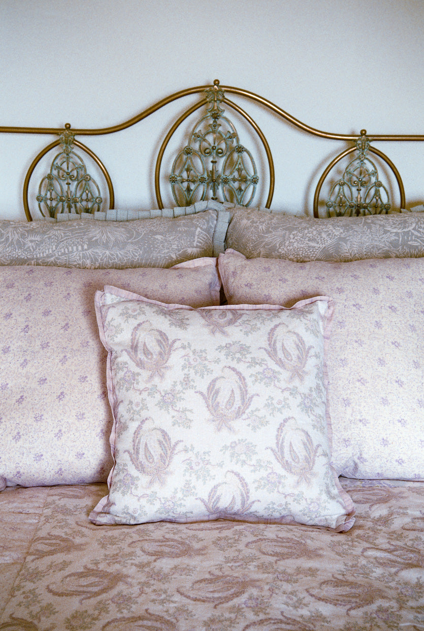 A close-up of a bed with a decorative gold metal headboard, featuring Floral Lavender, Paisley Stripe Lavender, and Storybook pillows, designed by Kathryn M. Ireland