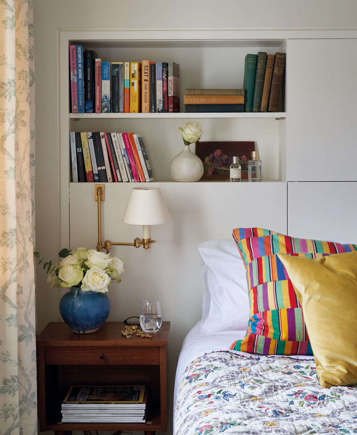 A cozy bedroom corner with a Froxfield Turquoise curtains, built-in white bookshelf filled with books, a brass wall lamp, a wooden nightstand with a vase of roses, and part of a bed, designed by Kathryn M. Ireland.