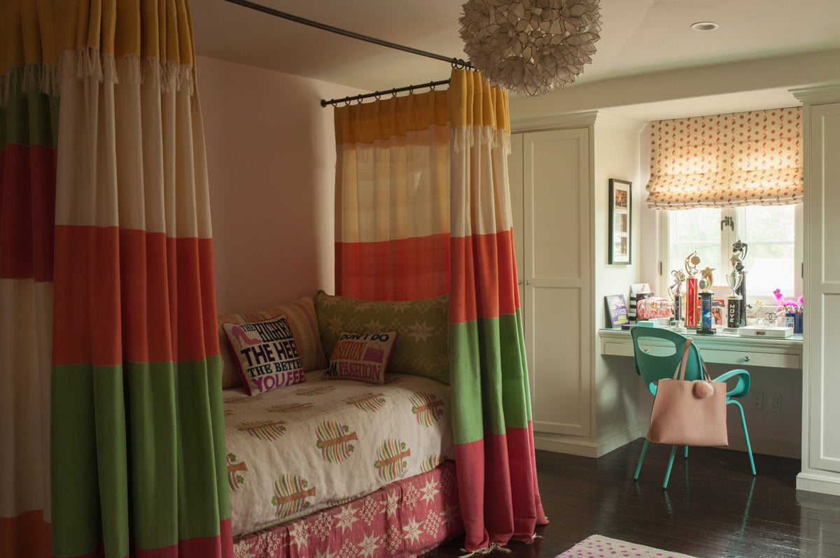 A bedroom scene with a bed dressed in a Fez Palm Calypso Orange bedding, a Gitana Margarita Green pillow, Gitana Red bed curtains, and Casablanca Natural Natural/Bubblegum Pink & Margarita Green roman shade, designed by Kathryn M. Ireland