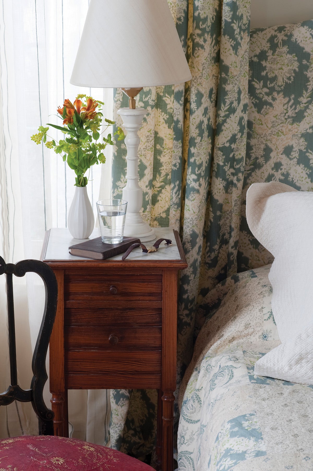 A vintage-style wooden nightstand in a bedroom, positioned next to a bed with Ikat Blue bedding, headboard, and curtains, designed by Kathryn M. Ireland