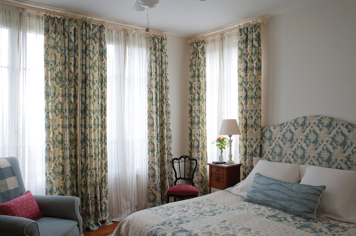 A styled bedroom featuring Ikat Blue curtains beige with Sheer Stripe Blue inner drapes, complemented by a matching patterned upholstered headboard and bedspread in Ikat Blue and a pillow in Moroccan Weave Blue, all designed by Kathryn M. Ireland.