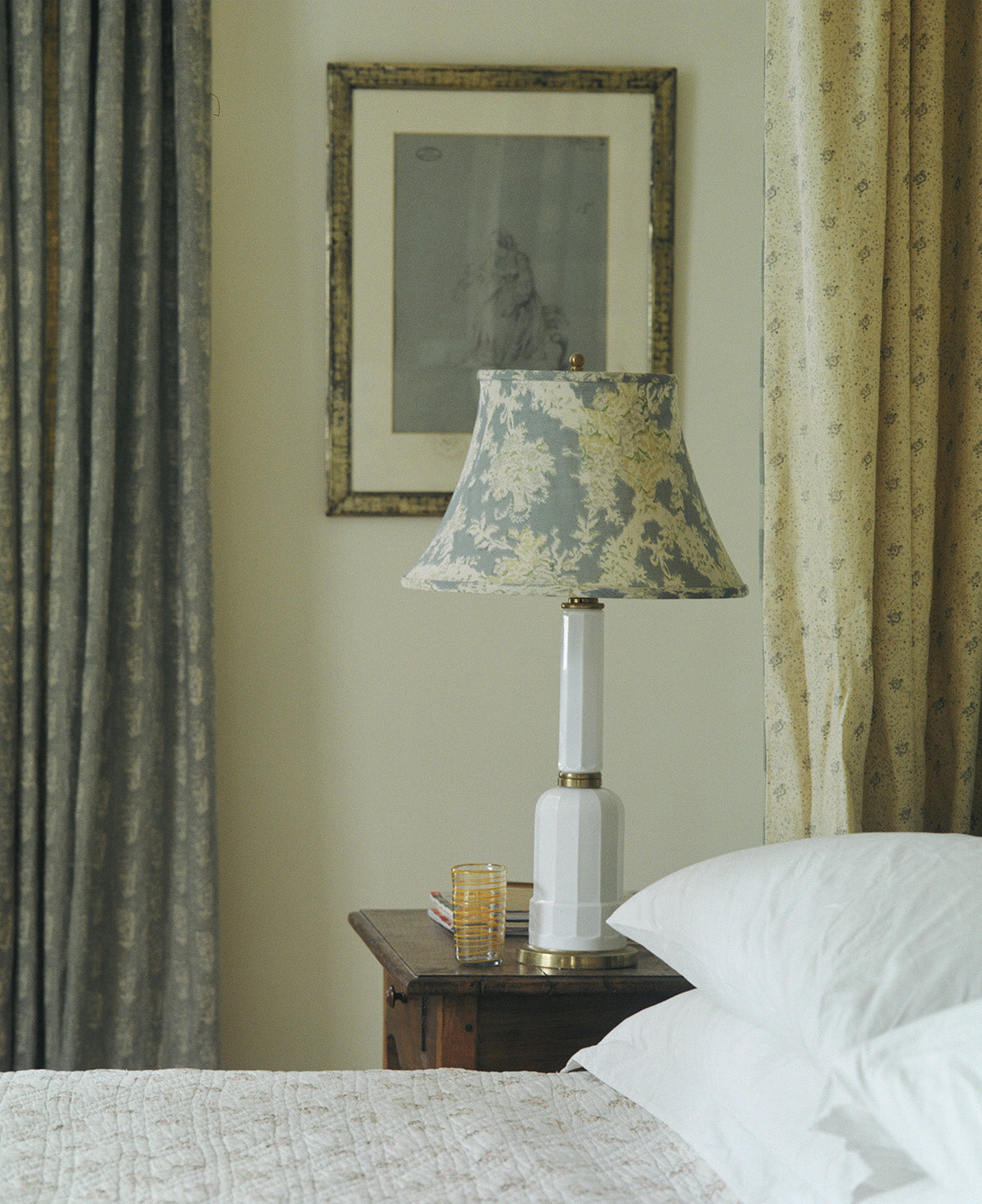A bedroom scene featuring a table lamp with an Ikat Blue lampshade, set on a dark wood nightstand beside a bed with white pillows, with Floral Blue curtains, designed by Kathryn M. Ireland