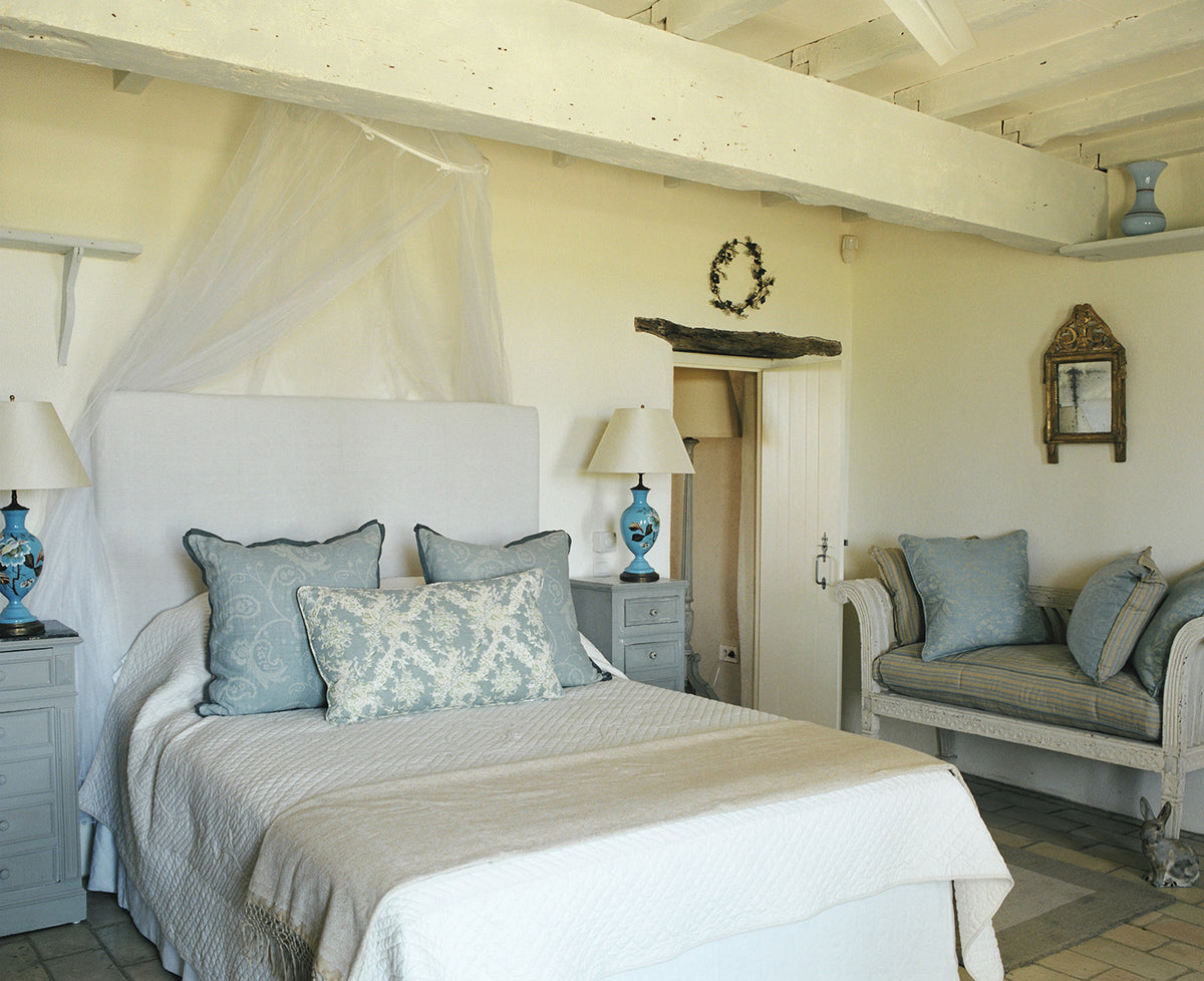 A rustic bedroom featuring a large bed with a white canopy, beige quilted bedding, an Ikat Blue pillow, set in a room with white wooden ceiling beams, matching bedside tables, and a matching armchair designed by Kathryn M. Ireland