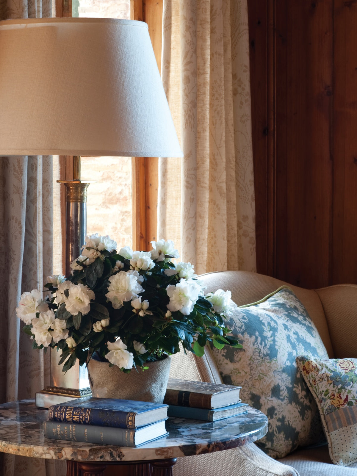 A styled interior scene with a table lamp, a vase of flowers, books on a marble side table, and a beige arm chair with an Ikat Blue pillow, designed by Kathryn M. Ireland