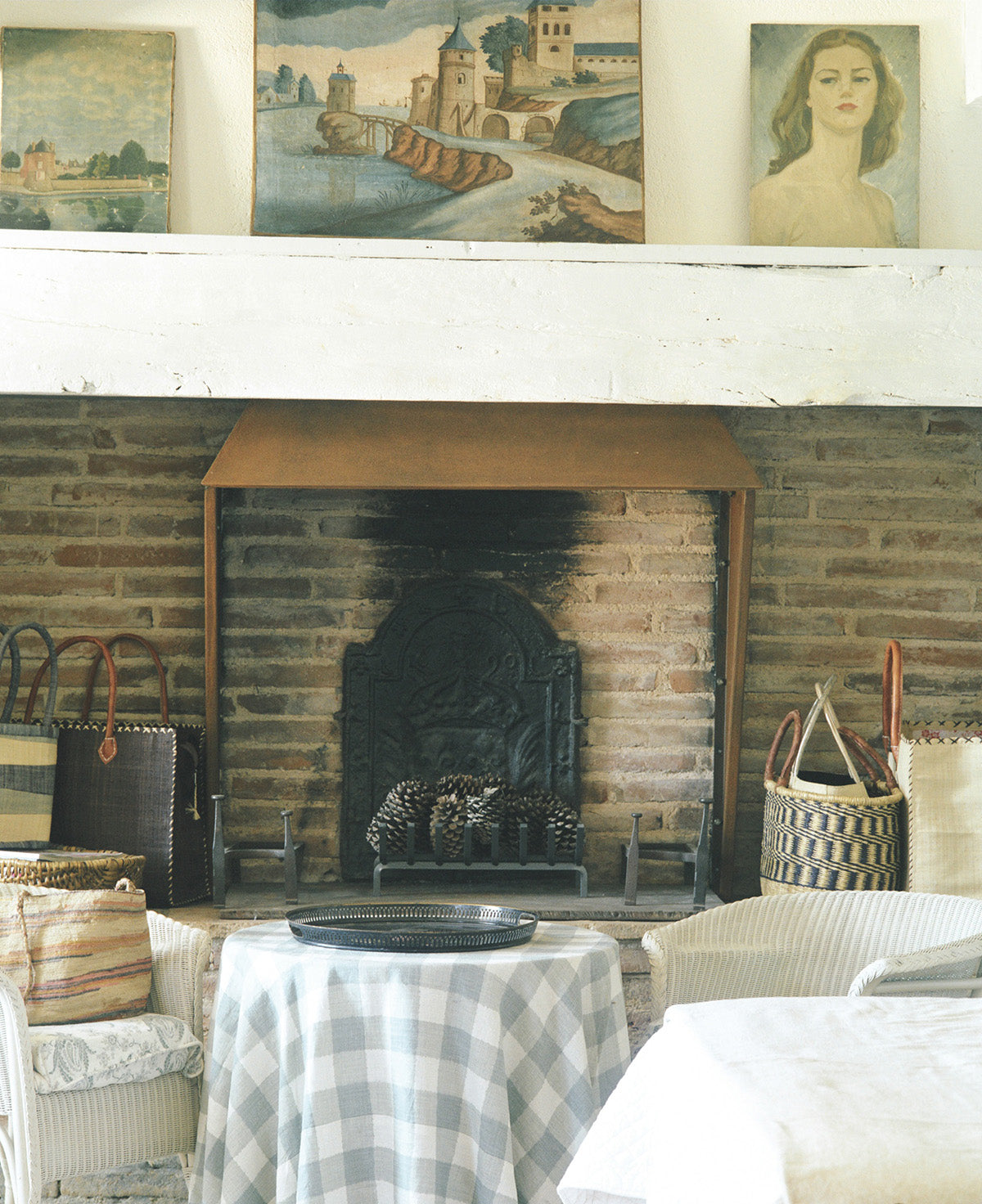 A rustic brick fireplace with a white mantel holding three framed paintings, with a small table covered in a Ikat Check Blue cloth designed by Kathryn M. Ireland
