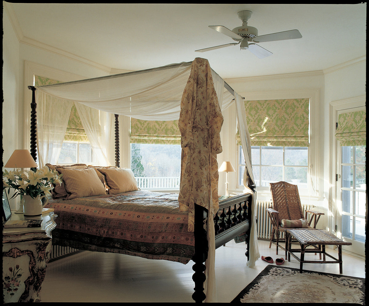 A classic four-poster canopy bed with a dark wood frame, dressed in beige and brown patterned bedding, set in a bright, traditional bedroom with Ikat Green window blinds, a ceiling fan, and a matching armchair designed by Kathryn M. Ireland