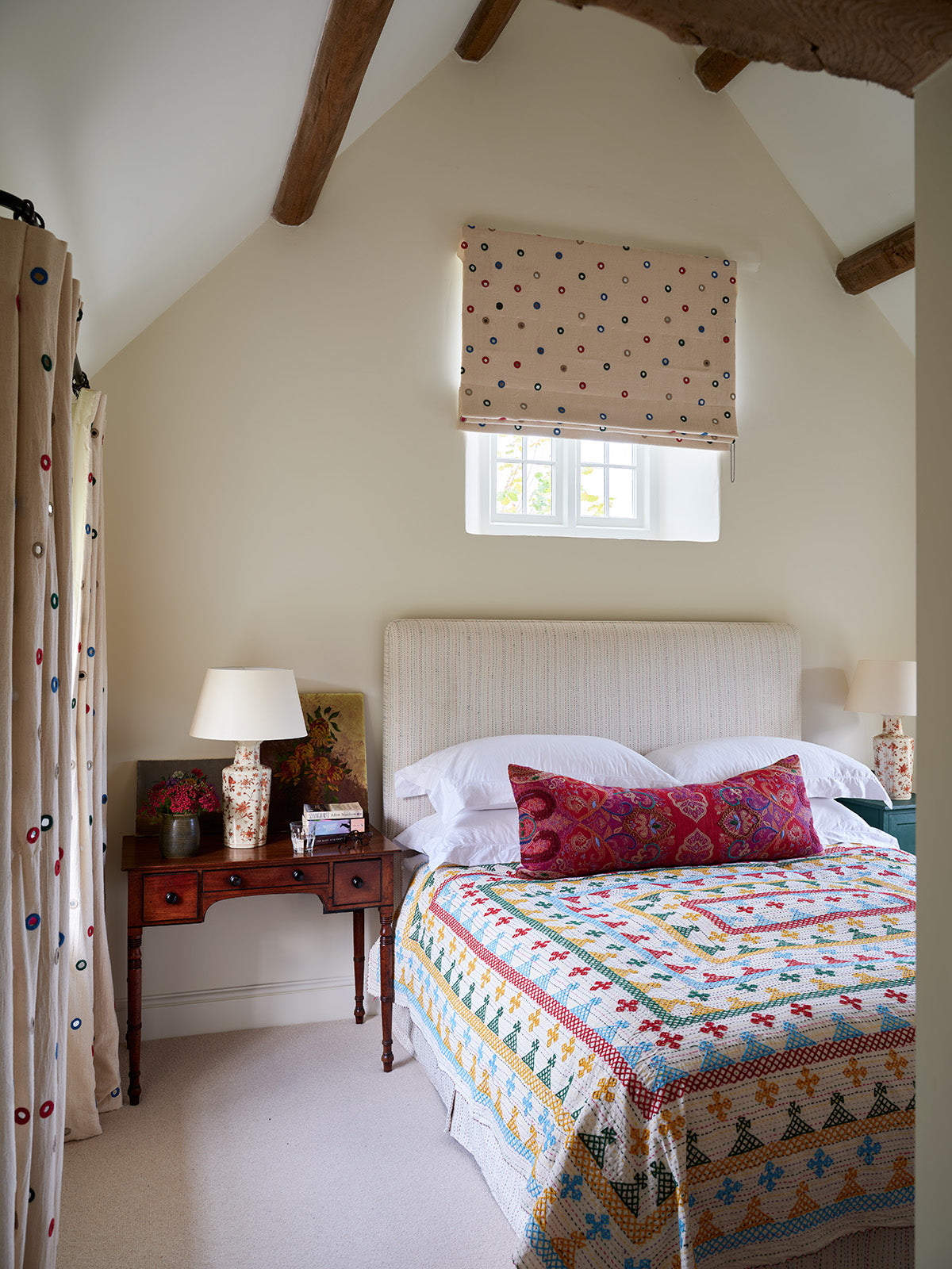 A bedroom interior featuring a bed dressed in a colorful geometric patterned quilt, and windows with Indian Mirror Multi roller blinds and curtains designed by Kathryn M. Ireland.