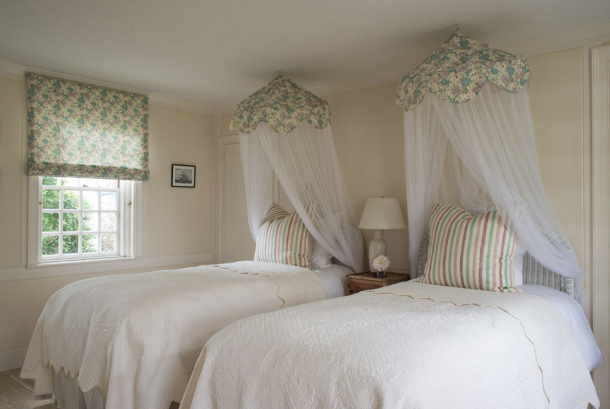 A cozy bedroom featuring two single beds with bedding and matching floral canopy bedsheets with sheer white canopies and pillows in Ithaca Mauve/Lavender/Pistachio, designed by Kathryn M. Ireland