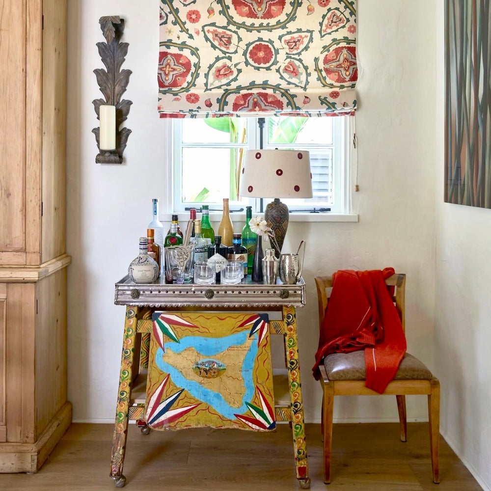 A bar cart in Kathryn M. Ireland's home with a lamp in Indian Mirror Maroon and a Bukhara Suzani Indigo window treatment.