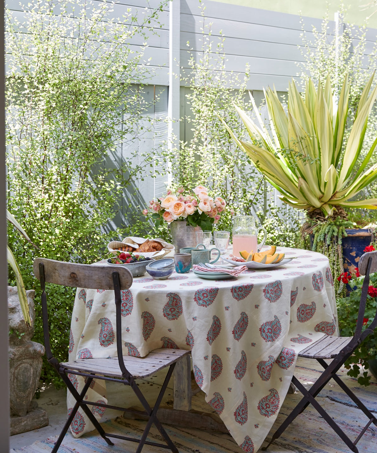 A round outdoor dining table set in a garden patio scene, covered with a tablecloth in Kirkbean Red (Turquoise), designed by Kathryn M. Ireland.