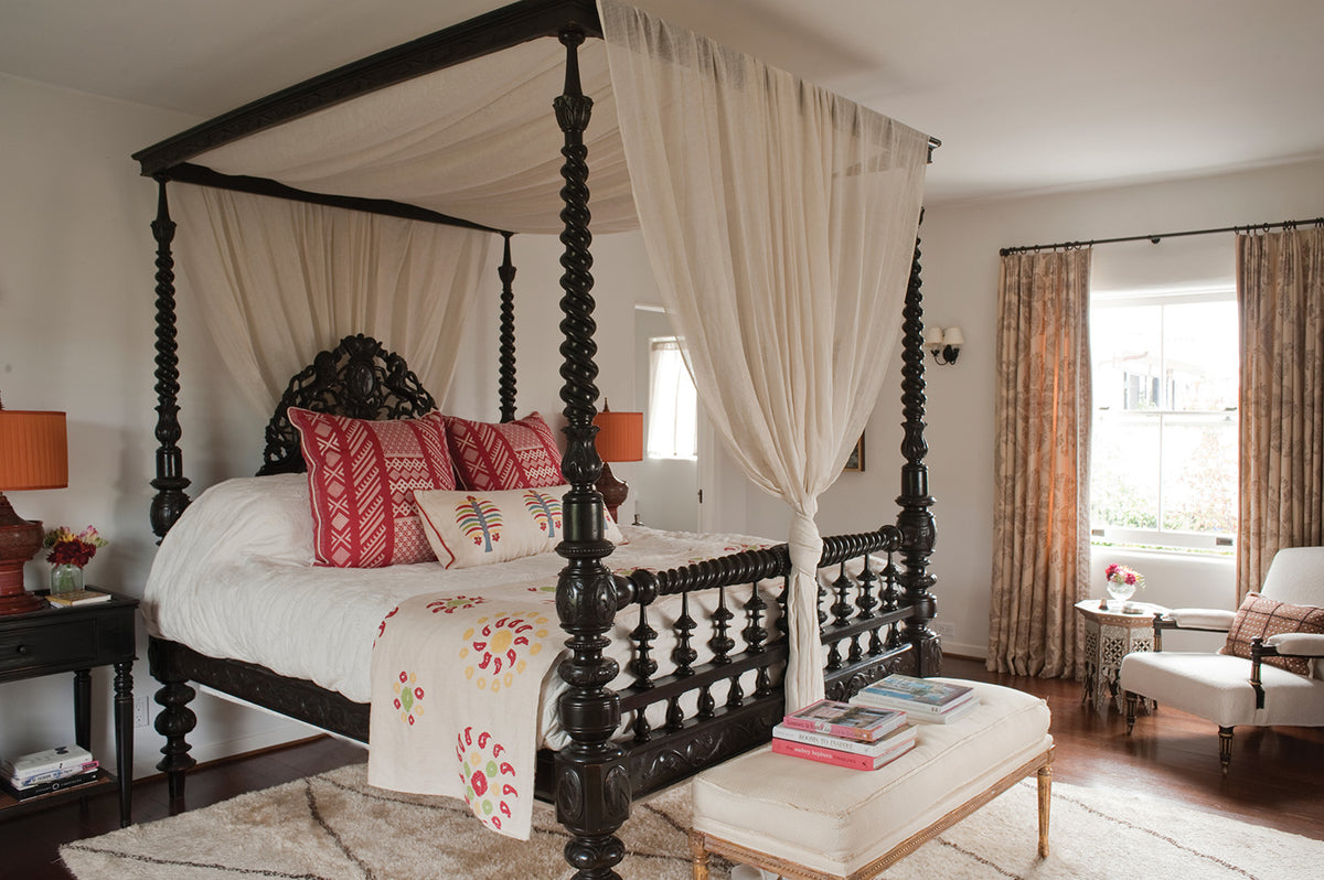 A classic four-poster canopy bed with a dark carved wooden frame, dressed in white bedding and pillows in Tangier Natural Cherry Red/Natural and Fez Palm Electric Blue, a Natural Marrakech Natural/Cherry Red bedspread, with Sheer Stripe White drapes, designed by Kathryn M. Ireland