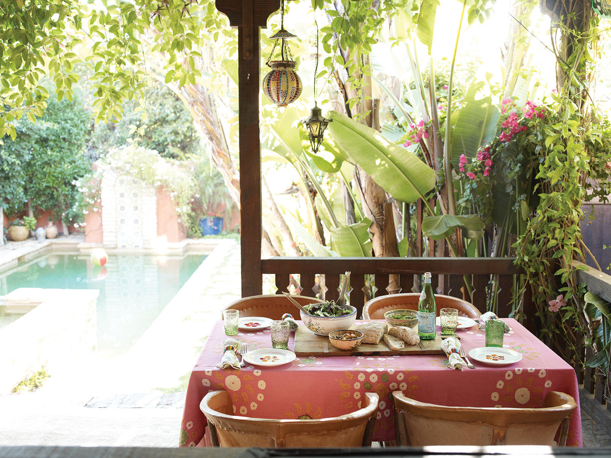 A rustic outdoor dining setup on a wooden pergola overlooking a tropical garden and swimming pool. A Marrakech Bubblegum Pink tablecloth covers a rectangular table set with plates, glasses, and green bottles, surrounded by woven chairs designed by Kathryn M. Ireland