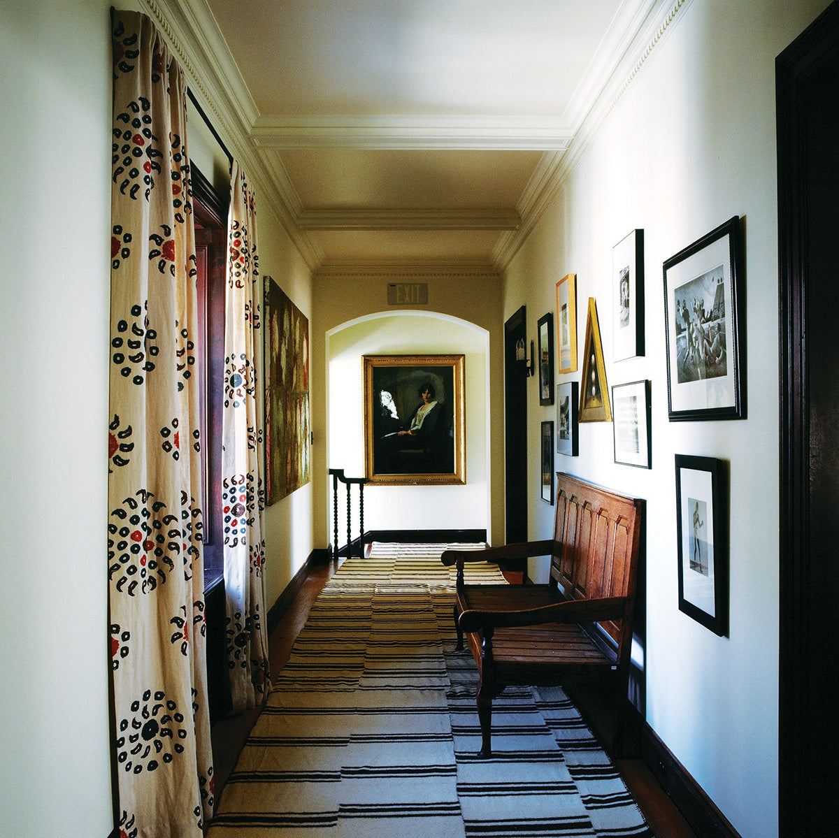 A long hallway with cream walls, framed black-and-white artwork, a wooden bench, Natural Marrakech Natural/Chocolate Brown curtains, and a striped rug leading toward a framed portrait at the end designed by Kathryn M. Ireland