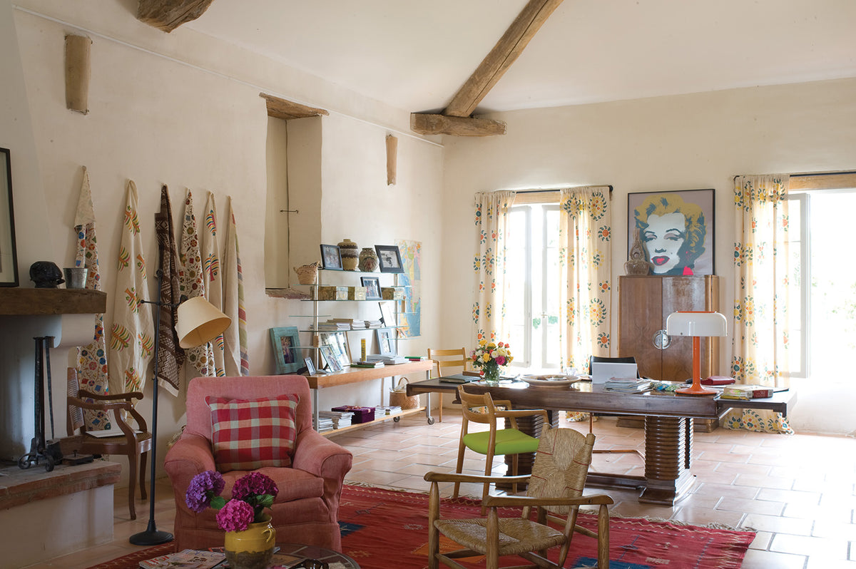 A rustic, cozy living room with exposed wooden ceiling beams, terracotta tile floors, and a pink armchair with a red plaid cushion in the foreground. The room features a wooden writing desk, Natural Marrakech Natural/Teal curtains, designed by Kathryn M. Ireland, and a colorful Marilyn Monroe portrait on the wall.