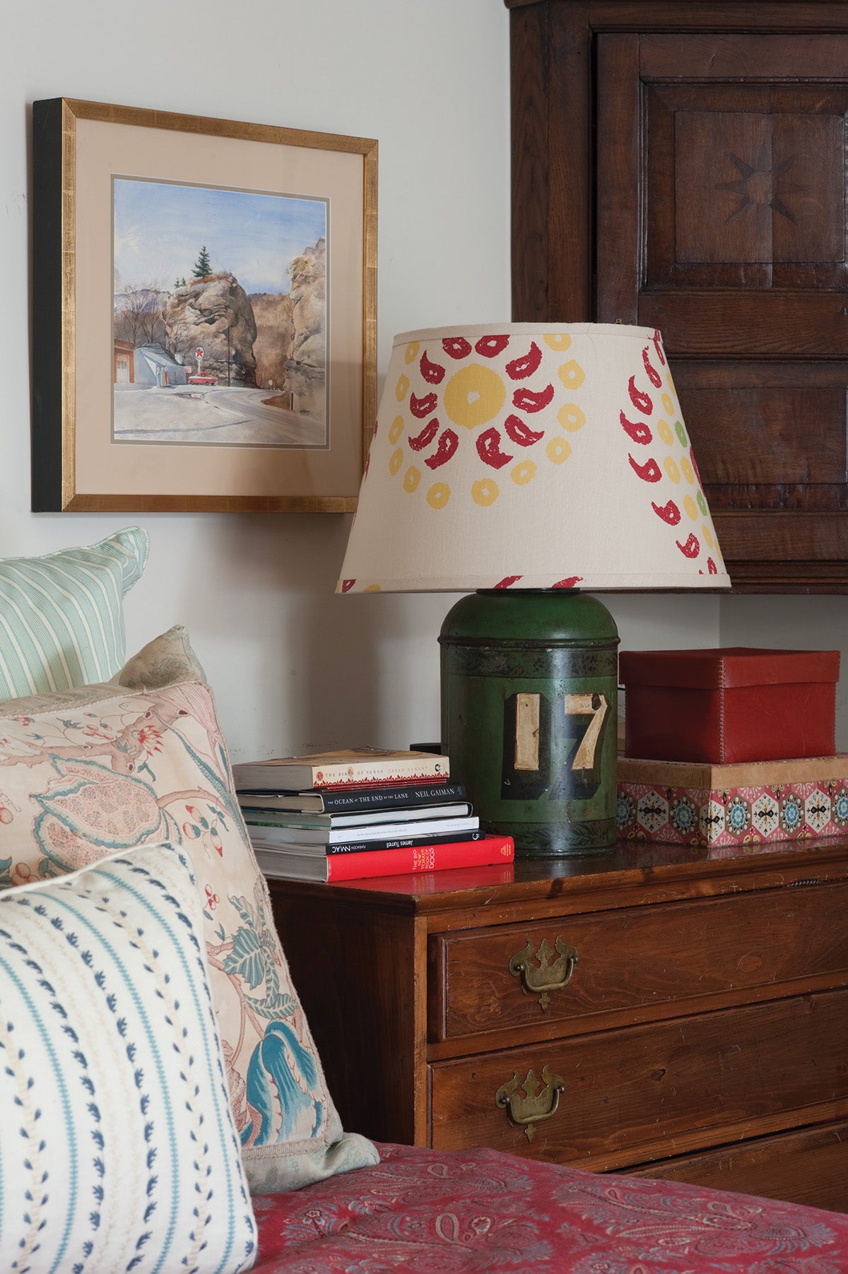 A styled bedroom corner featuring a Natural Marrakech Natural/Cherry Red lampshade, a green metal lamp base, a wooden nightstand, and a framed landscape print on the wall, designed by Kathryn M. Ireland