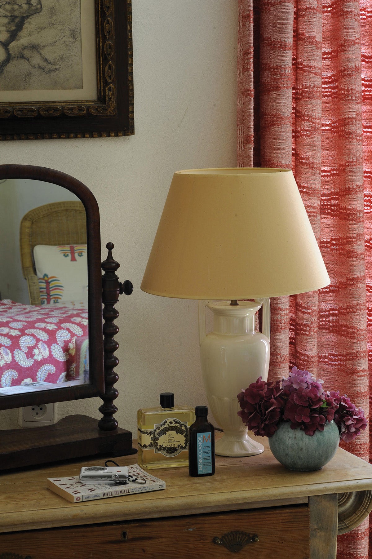 A beige table lamp with a cream ceramic base sits on a wooden dresser next to a mirror with Moroccan Weave Red curtains designed by Kathryn M. Ireland
