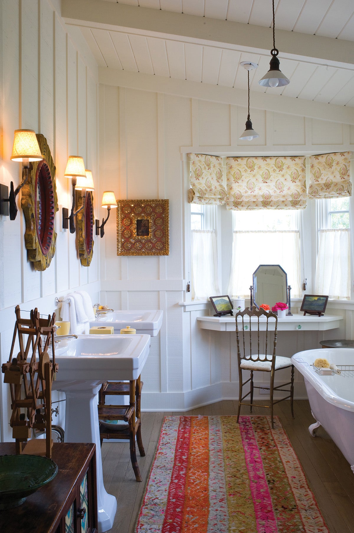 A bright bathroom interior with white paneled walls, double white pedestal sinks, wall-mounted lamps, a window with Paisley Stripe Red curtains, a vanity desk, and a colorful striped runner rug leading toward a white clawfoot bathtub, designed by Kathryn M. Ireland.