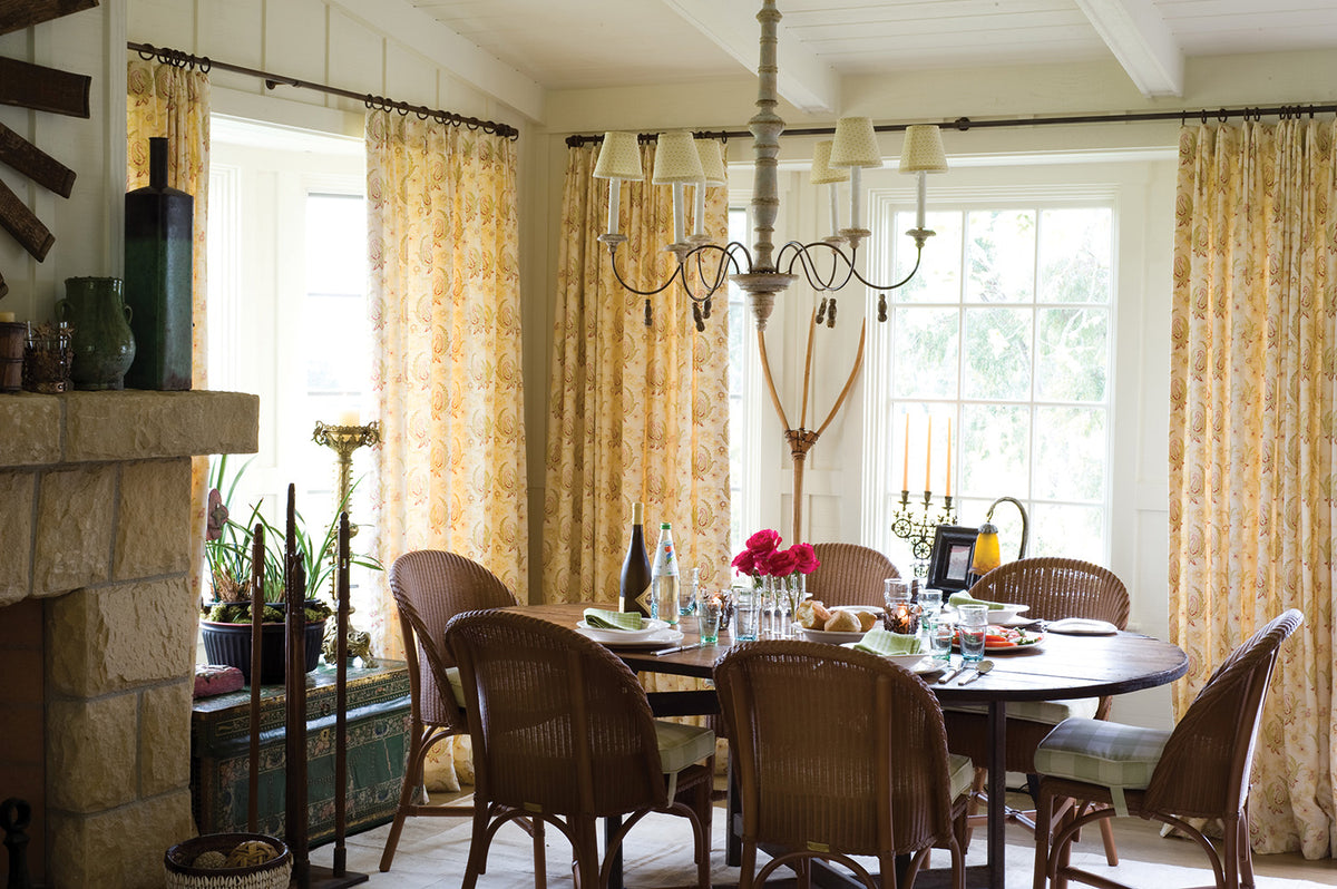 A dining room interior with a round wooden table, wicker chairs, a hanging chandelier, and tall Paisley Stripe Red curtains around large windows designed by Kathryn M. Ireland