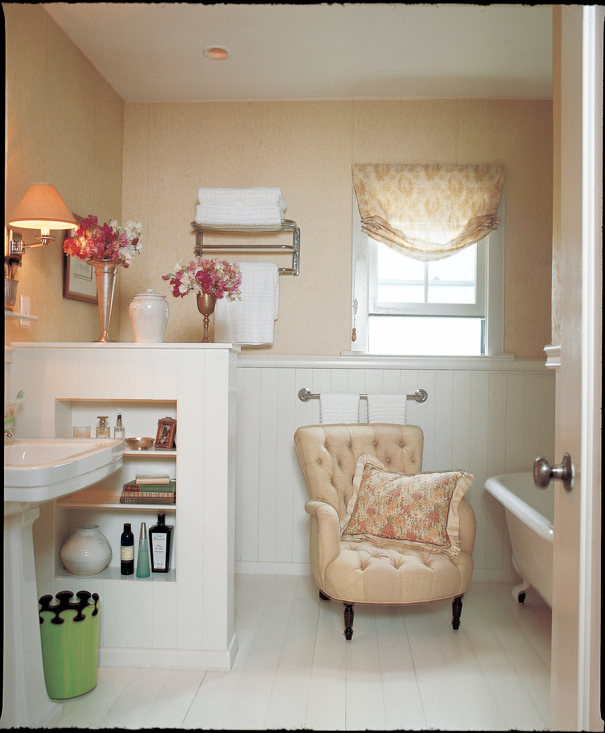 A bright bathroom interior featuring a tufted cream armchair with a floral accent pillow, a white pedestal sink, white wainscoting, and a window with a Paisley Stripe Red curtain designed by Kathryn M. Ireland
