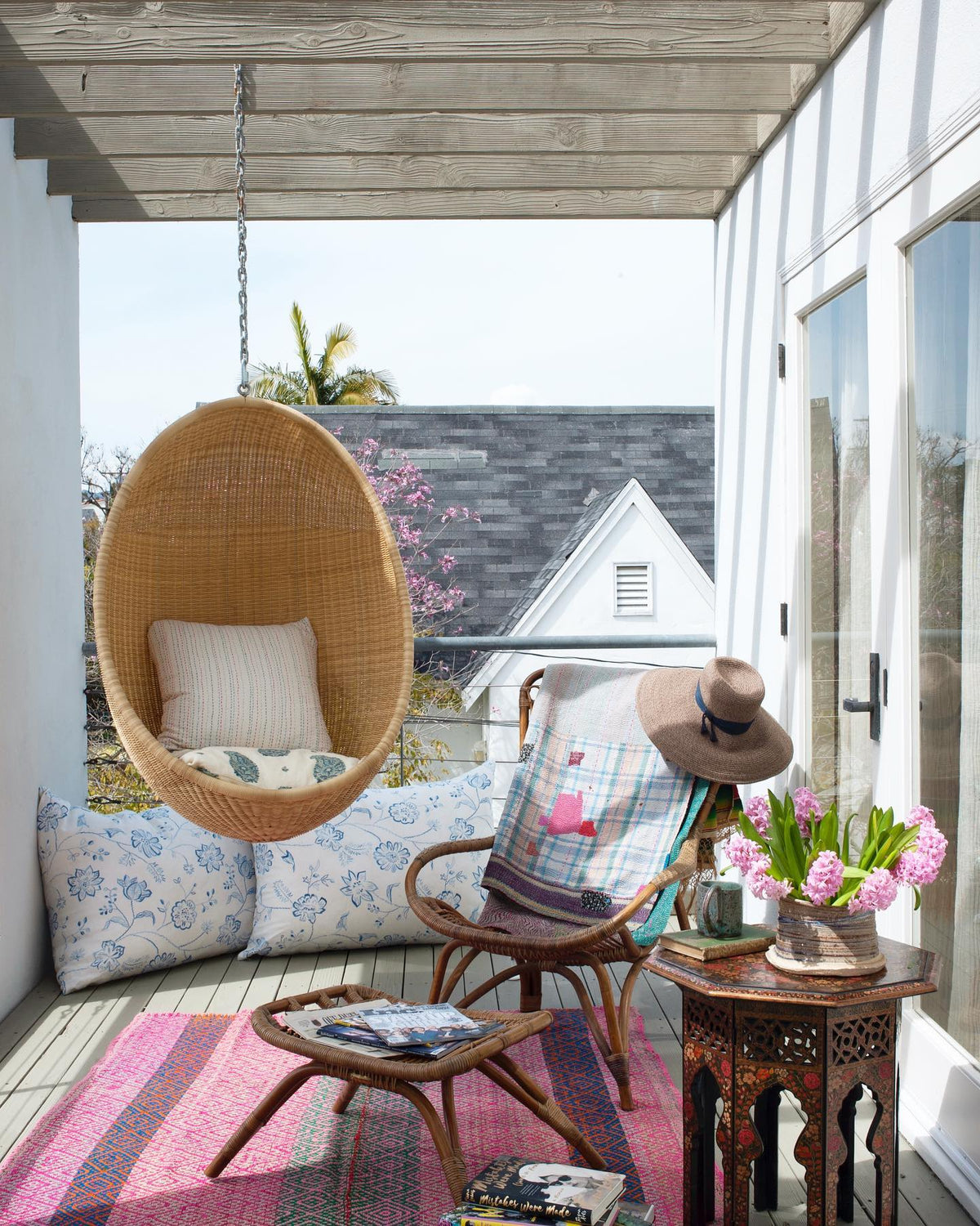 A styled outdoor patio seating area featuring a woven hanging egg chair with pillows in Kirkbean Turquoise (Blue) and Hand-Stitched Multi, a wooden chair with a plaid throw and a wide-brim hat, and large pillows in Northhaven Blue, designed by Kathryn M. Ireland.