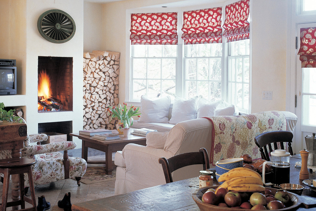 A cozy living room interior with a cream sofa, patterned armchair, fireplace, stacked firewood, and Quilt Red window valances designed by Kathryn M. Ireland