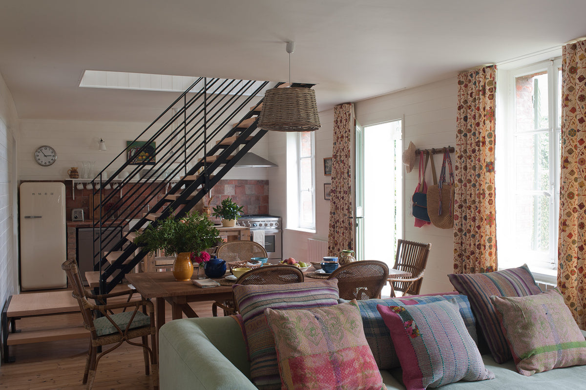 A cozy, rustic-style home interior featuring a light green sofa, a wooden dining table, a black metal staircase, and Safi Suzani Autumn curtains by tall windows designed by Kathryn M. Ireland
