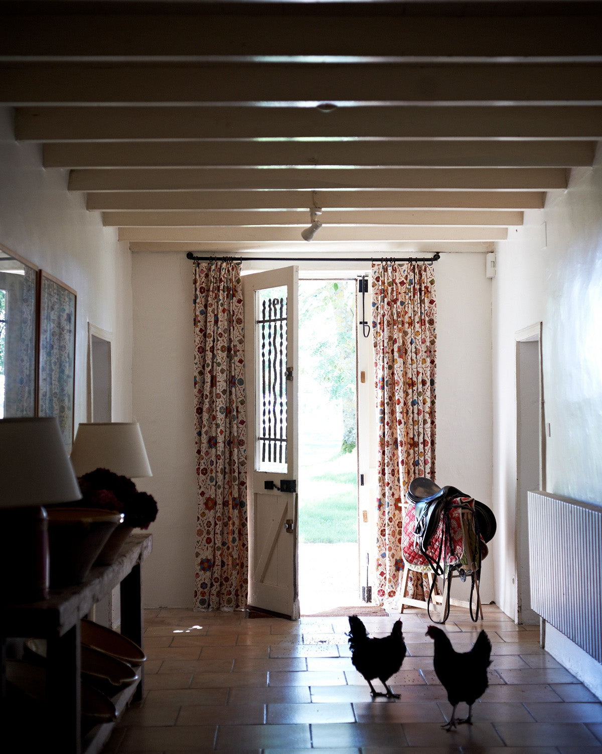 A long interior hallway with exposed ceiling beams, Safi Suzani Autumn curtains framing an open doorway, and two black chickens walking on a tiled floor, designed by Kathryn M. Ireland
