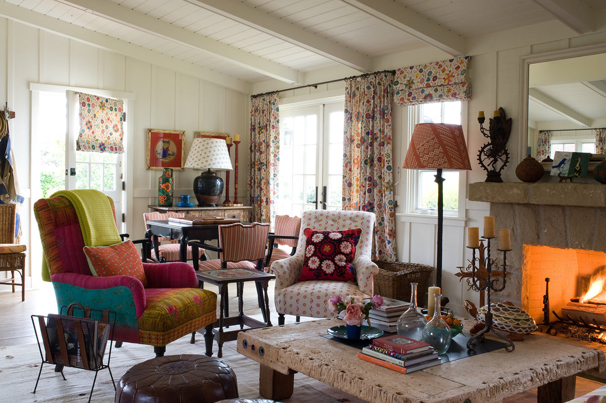 A cozy, rustic living room with a colorful accent chair, a Casablanca Bubblegum/Sunburst armchair, a rustic stone coffee table, Safi Suzani Fiesta curtains, and a warm fireplace in the background, designed by Kathryn M. Ireland