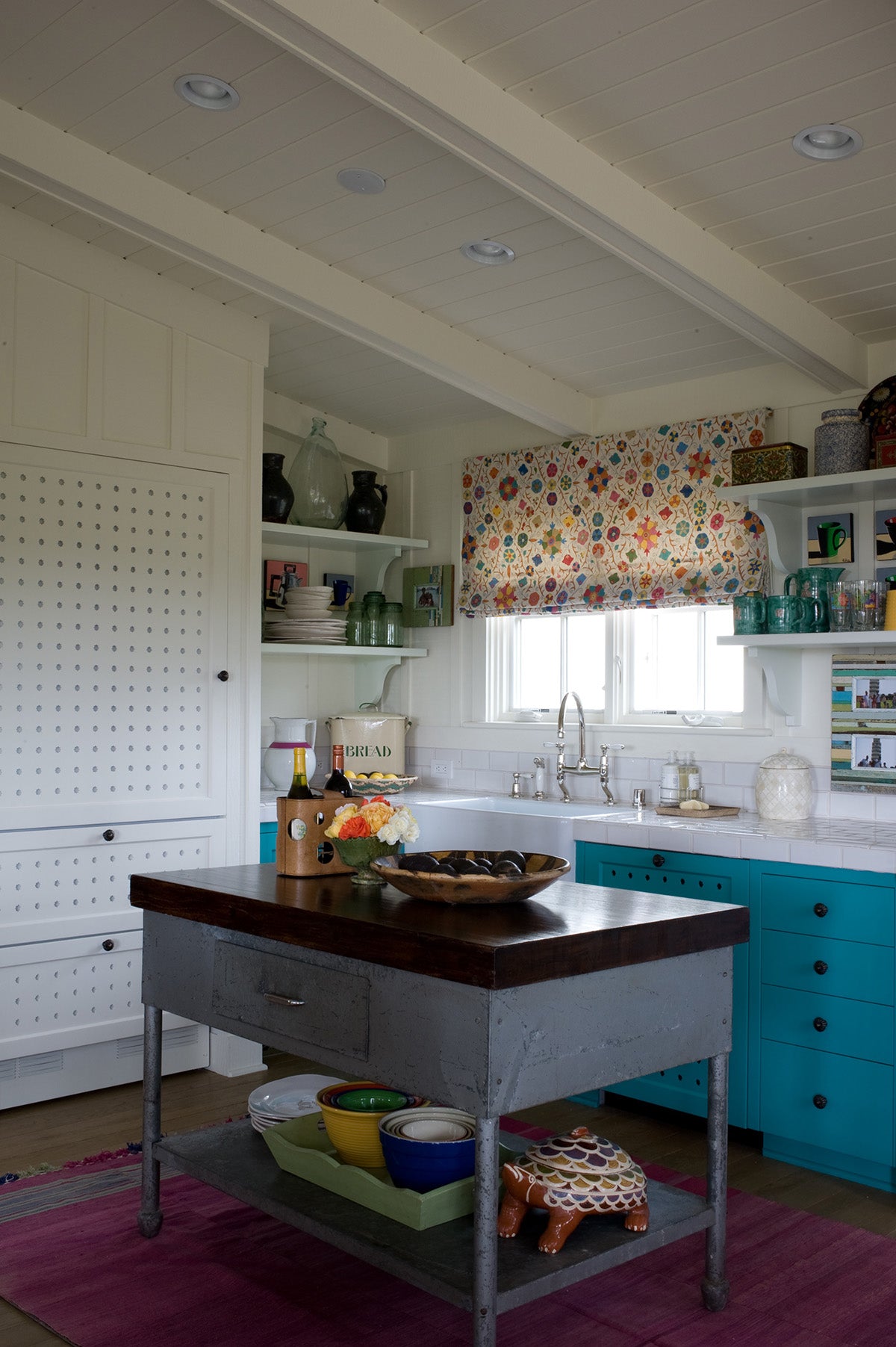 A bright kitchen interior with a central island table, turquoise cabinets, white cabinetry, a Safi Suzani Fiesta window valance, and colorful bowls and dishes on the island, designed by Kathryn M. Ireland