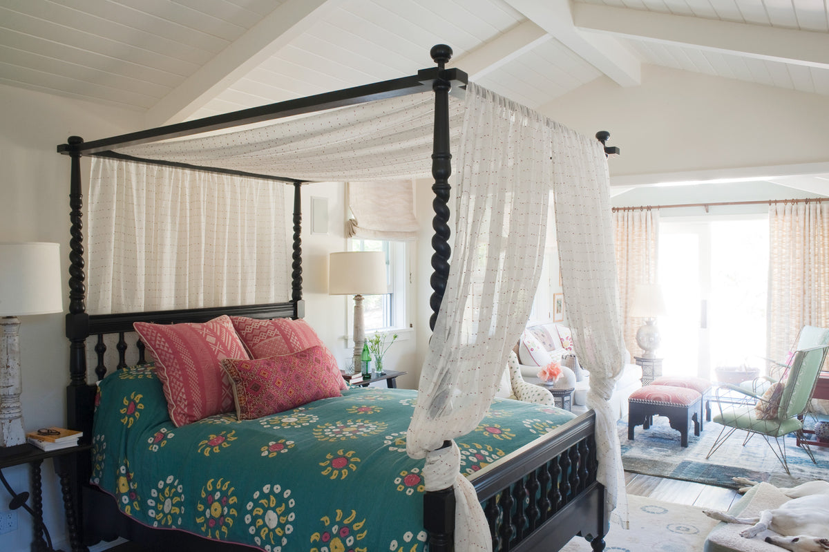 A black wooden four-poster canopy bed with Sheer Confetti Desert Rose curtains, Marrakech Teal bed spread, and Tangier Natural Cherry Red/Natural pillows designed by Kathryn M. Ireland