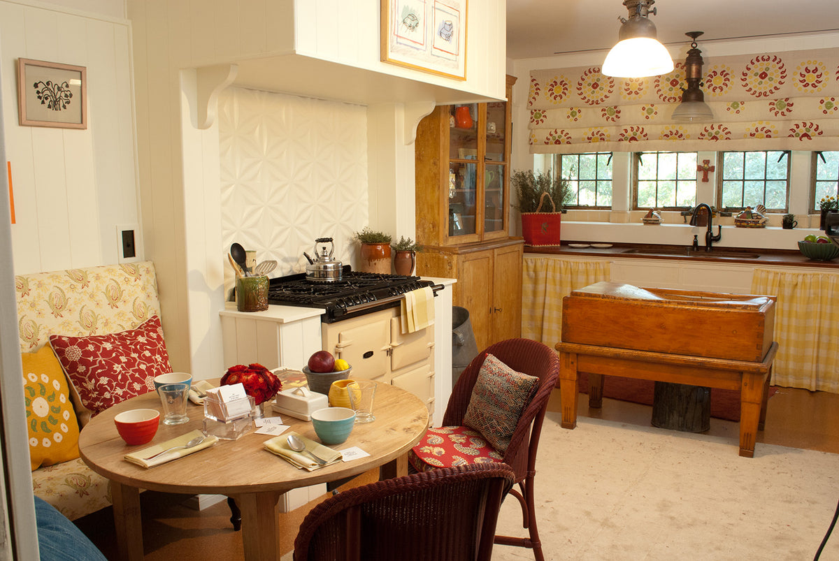 A cozy, vintage-style kitchen and dining area with warm wood cabinetry, a small round wooden table set with dishes, a Paisley Stripe Red built-in bench, an Abu Red Currant pillow and a Marrakech Tangelo pillow, Small Check Yellow curtains below the sink and Marrakech Natural Cherry Red curtains above the window designed by Kathryn M. Ireland