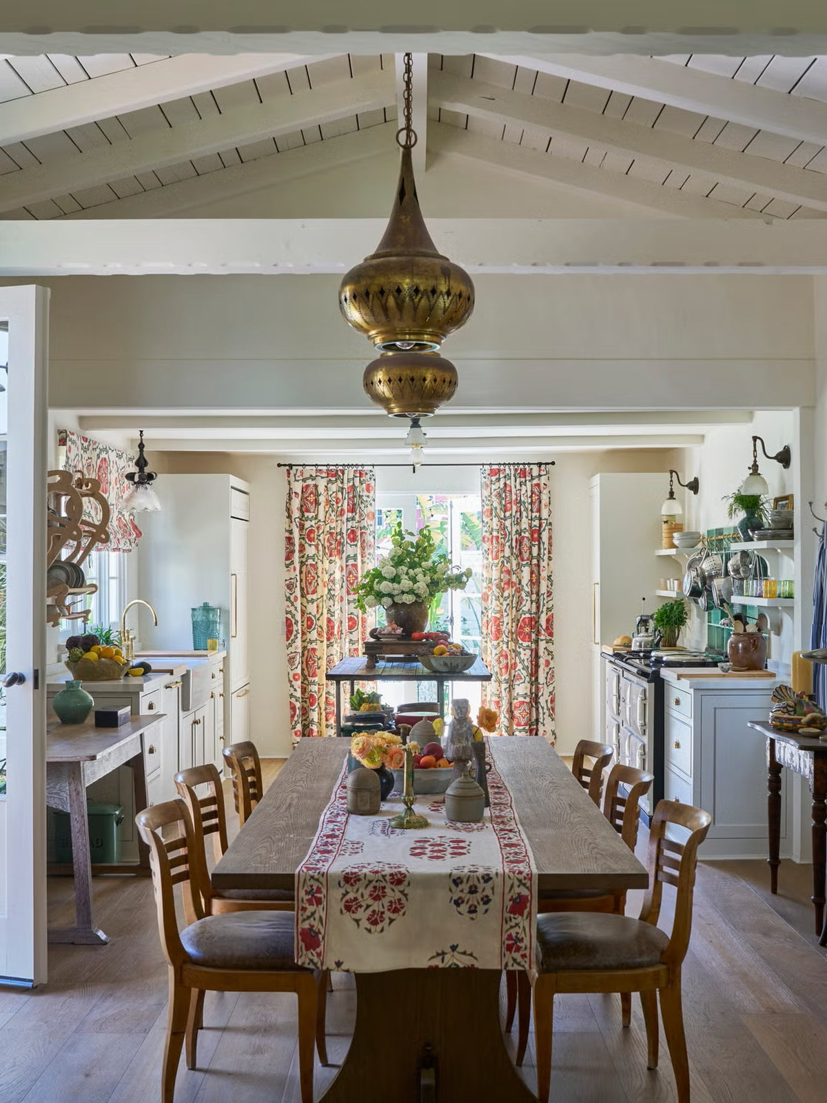 A large brass pendant light hangs over a dining table in a bright, traditional kitchen and dining room with Sultan Suzani Orange curtains and Hilltop Paisley Regal table runner, designed by Kathryn M. Ireland.