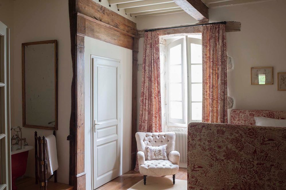 A cozy, rustic bedroom interior featuring a vintage tufted armchair, a bed in Toile Red  bedding, and matching curtains by a bright window, framed by exposed wooden beams and a mirror, designed by Kathryn M. Ireland.