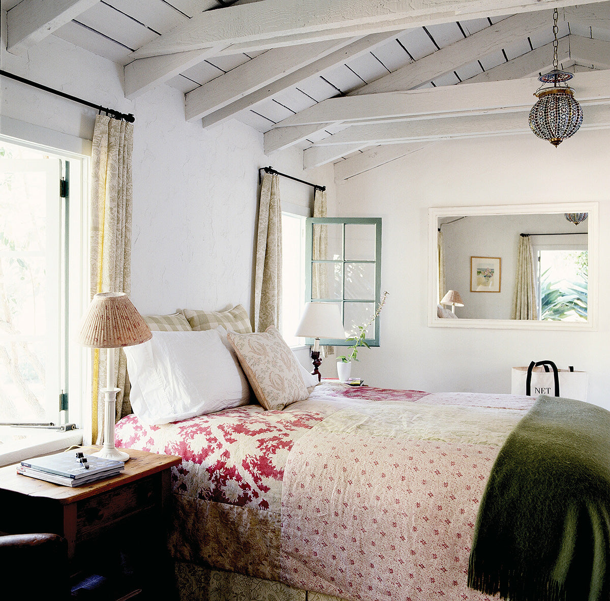 A rustic bedroom interior with Toile Silvertree curtains, a patchwork quilt including Ikat Red and Floral Red fabric, pillows in Ikat Check Green, designed by Kathryn M. Ireland