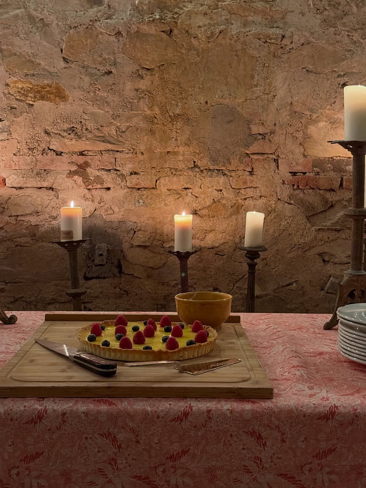A fruit tart topped with fresh raspberries sits on a wooden board on a table covered with a Wildflowers Pink tablecloth, designed by Kathryn M. Ireland.